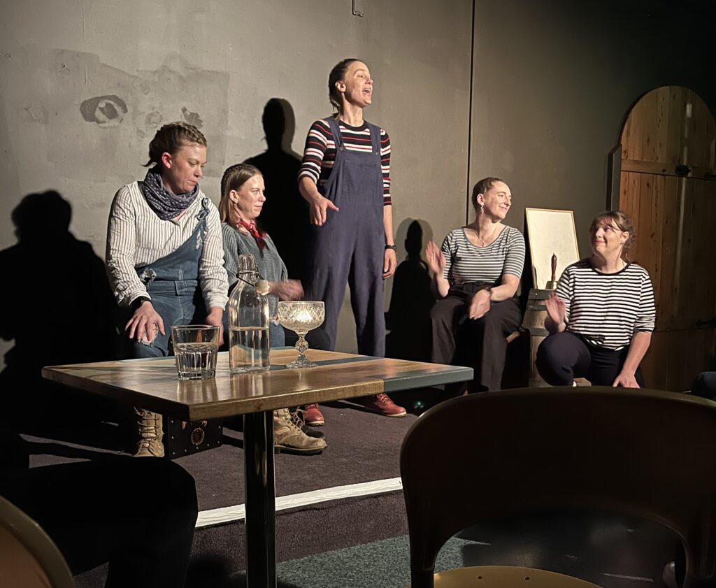 Five women with stripy shirts singing on a stage in a small pub space