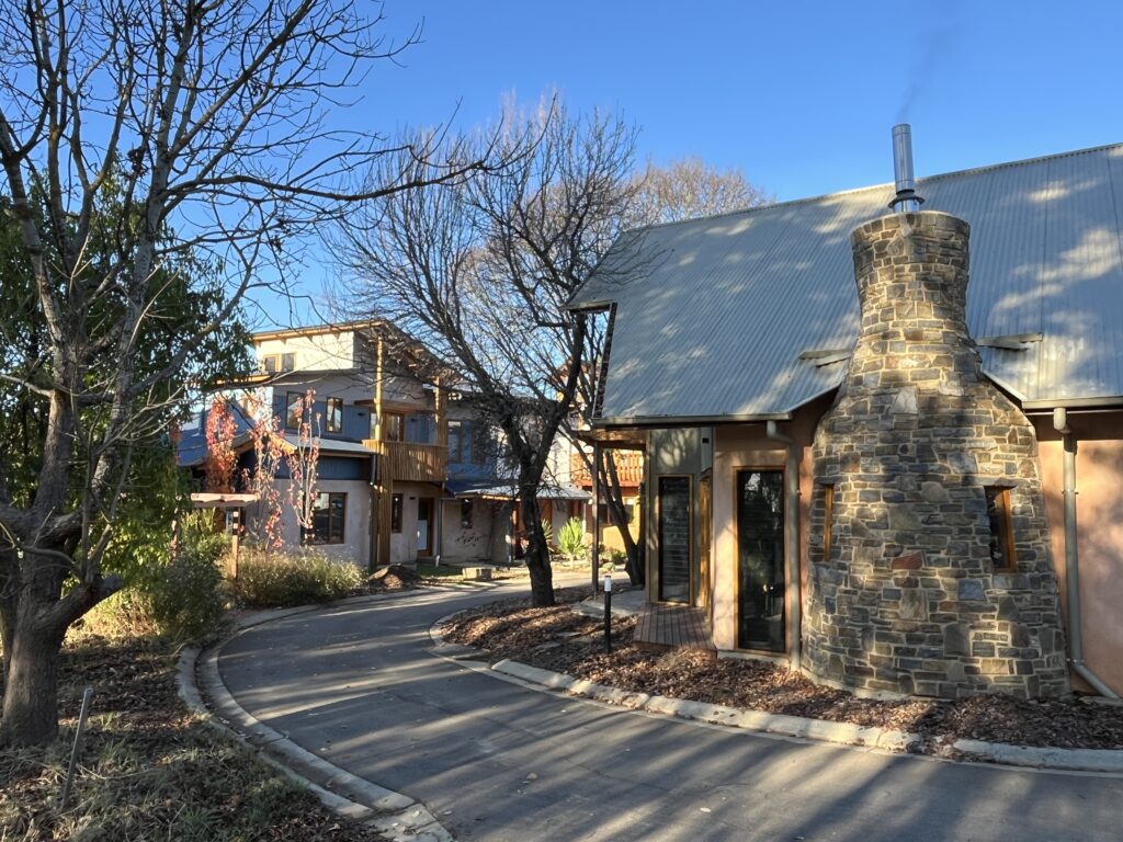 Long winter shadows of trees fall on an earthy building with a circular stone chimney. There is a narrow road around it, with a two storey dwelling in the background.