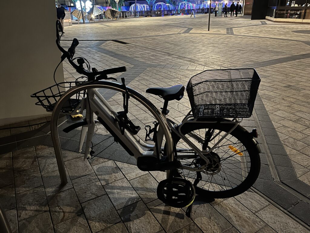 An ebike locked to a biker rack on a paved concourse at night. The front wheel is missing