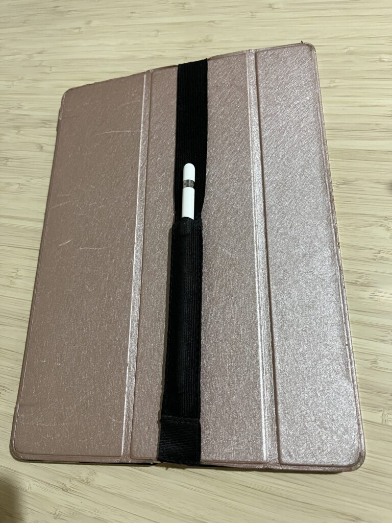 A pink iPad cover with a piece of black elastic with a pouch in it strapped to the front holding an Apple Pencil to the front.