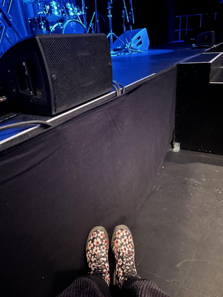 The front of a stage bathed in blue light. At the bottom of the image, touching the stage, white Doc Martens with red flowers can be seen.