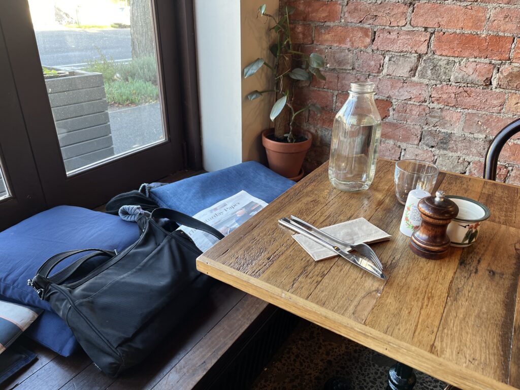 The corner table in a cafe next to a window sea with blue cushions. The brick wall is exposed, the table wooden and the servingware mismatched. There is a potplant in one corner and a handbag, gloves and a newspaper on the seat.