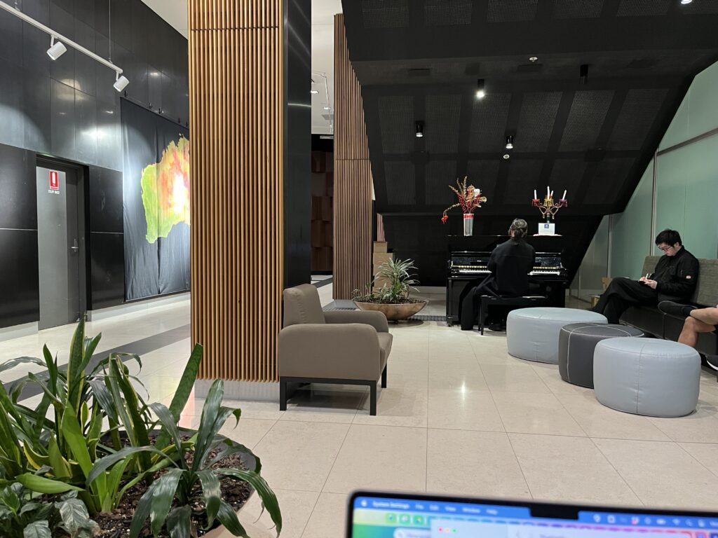 The lobby of a building. There is a woman under the stairs playing a piano and a 2 metre high map of Australia on one wall