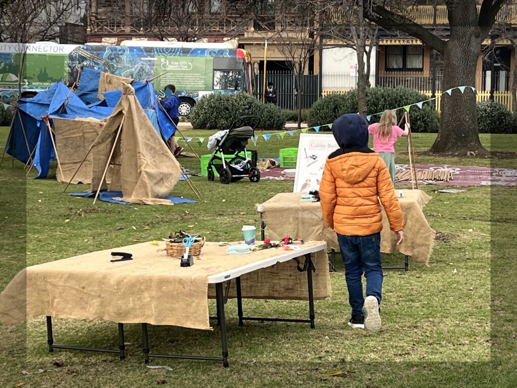 Two craft tables covered in hessian. Children are building cubbies with tarpaulins, hessian and wooden poles on the grass in the background
