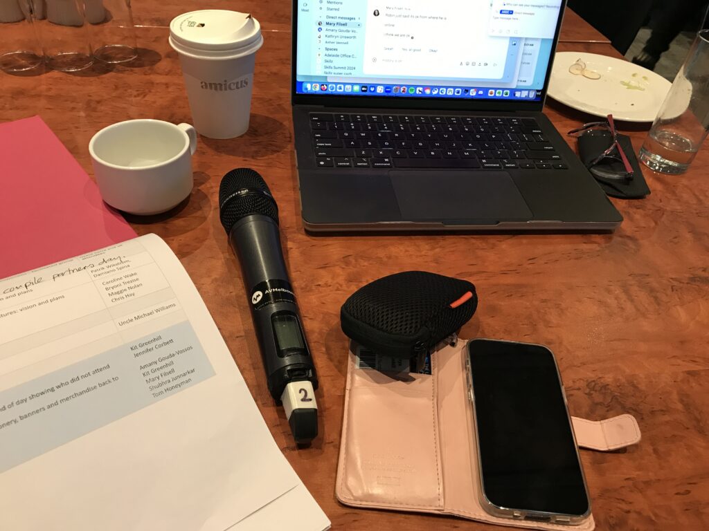 A table with a run sheet, microphone, two coffee cups, a drinking glass, iPhone, mouse glasses and MacBook.